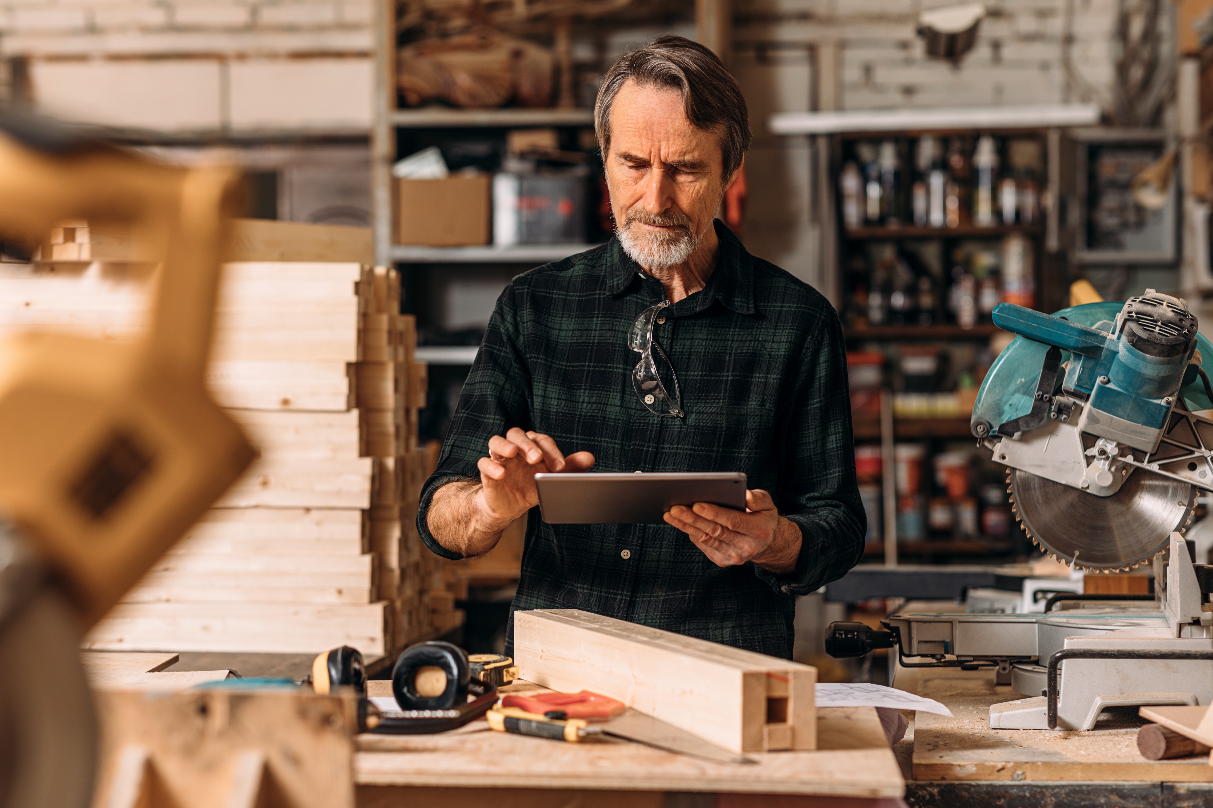A business owner in a woodworking shop reviewing work on a tablet, surrounded by lumber and tools
